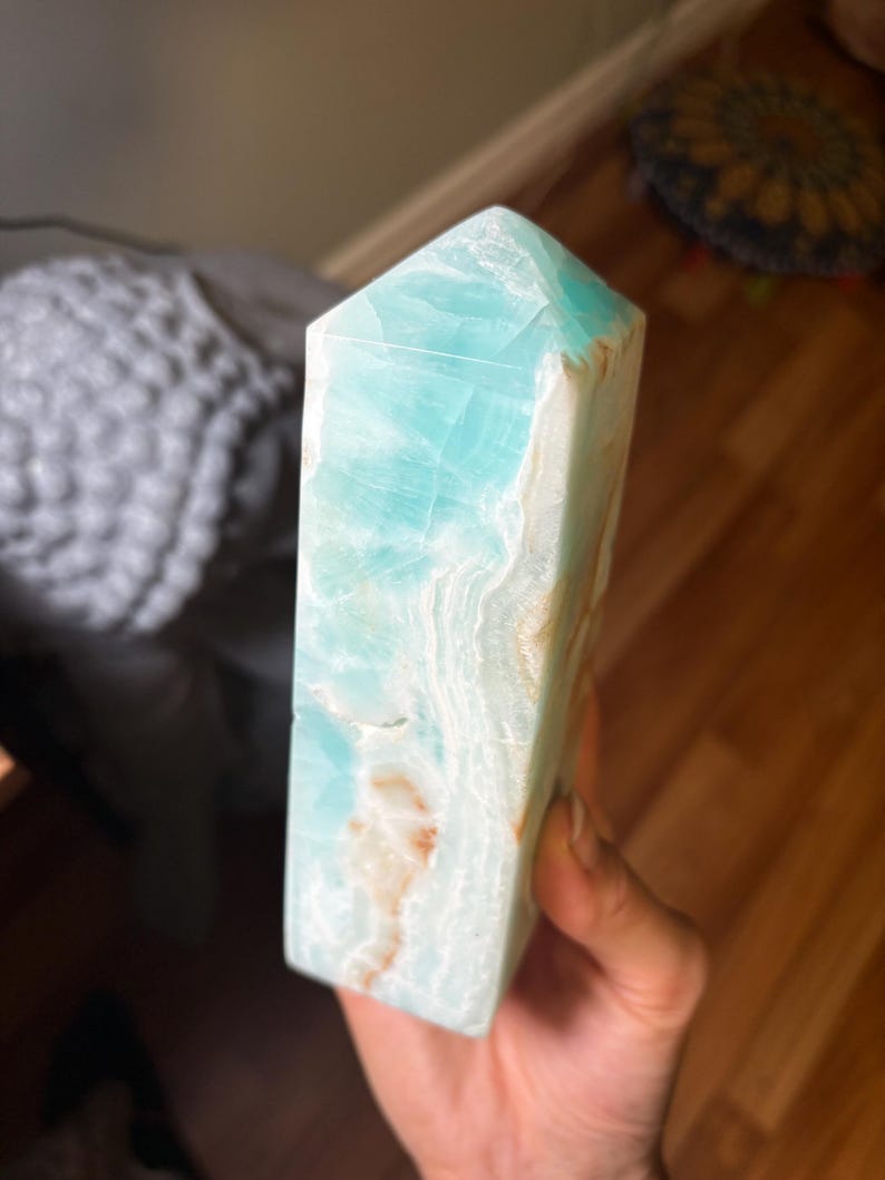 May include: A polished Caribbean calcite crystal tower featuring a hexagonal point. The stone displays a vibrant turquoise blue color with translucent layers and natural white and tan mineral inclusions running throughout the vertical structure.