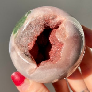 May include: A pink and white geode sphere with a large opening revealing a red and brown crystal interior. The sphere has a green and white pattern on the outside.