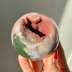 May include: A pink, green, and red agate sphere with a natural druzy geode opening. The sphere is smooth and polished, with a natural, organic shape.