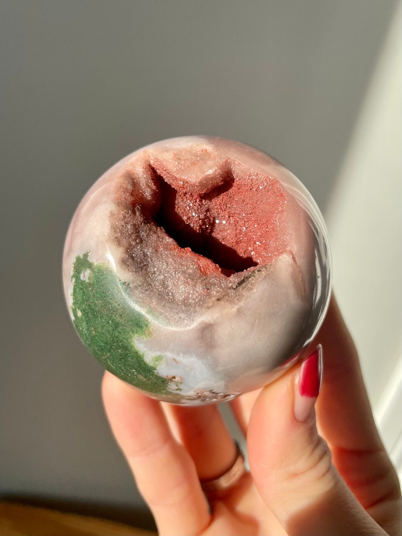 May include: A pink, green, and red agate sphere with a natural druzy geode opening. The sphere is smooth and polished, with a natural, organic shape.