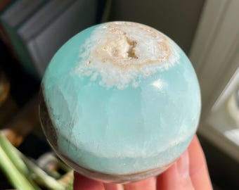 May include: A polished sphere of blue-green aragonite, with a band of tan and white near the top. The gemstone is held in a hand, with a blurred background of plants and a window.