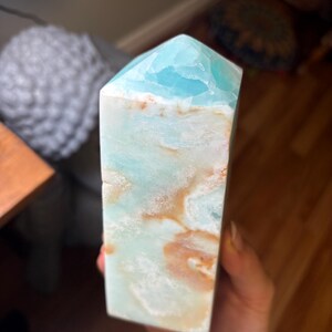 May include: A polished crystal tower with a hexagonal point, featuring a smooth surface with a marbled pattern of light blue, white, and tan colors. The stone has a translucent quality near the top and opaque sections throughout the body.