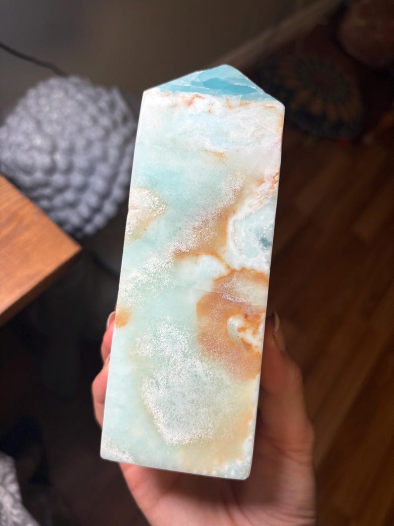 May include: A polished stone tower with a hexagonal point, featuring a smooth surface with a marbled pattern of light blue, white, and tan brown colors. The crystal structure displays translucent areas and opaque mineral inclusions throughout the piece.