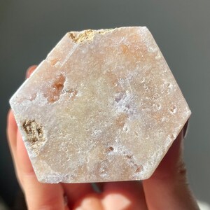 May include: A hexagonal, light pink and white selenite crystal with a rough, natural texture. The crystal has a translucent appearance and is slightly cloudy in color.
