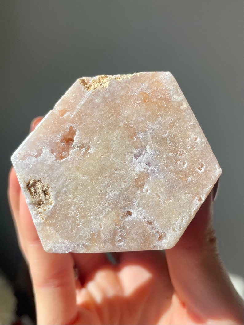 May include: A hexagonal, light pink and white selenite crystal with a rough, natural texture. The crystal has a translucent appearance and is slightly cloudy in color.
