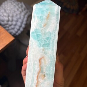 May include: A polished, tower-shaped crystal point featuring a translucent light blue and white banded pattern with distinct, thin, irregular orange-brown mineral inclusions running vertically through the stone's smooth, faceted surface.