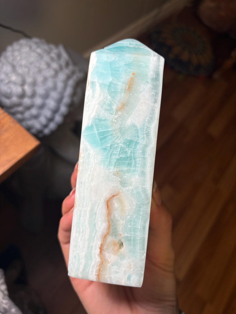 May include: A polished, tower-shaped crystal point featuring a translucent light blue and white banded pattern with distinct, thin, irregular orange-brown mineral inclusions running vertically through the stone's smooth, faceted surface.