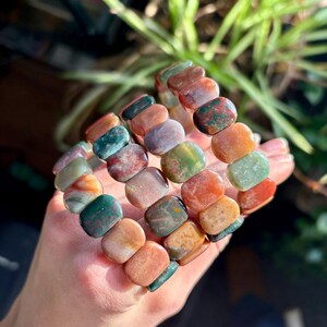 May include: Three gemstone bracelets with a variety of colors including green, orange, red, and purple. The bracelets are made of polished, rectangular stones. The bracelets are held in a hand.