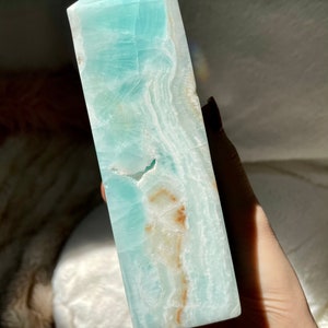 May include: A polished Caribbean calcite tower featuring a smooth, hexagonal point. The stone displays a vibrant light blue and teal color palette with translucent layers, white banding, and distinct patches of warm, earthy brown mineral inclusions.