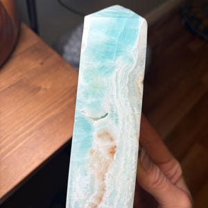 May include: A polished tower of Caribbean calcite featuring a vibrant light blue color with white, wavy, translucent banding and small patches of orange-brown mineral inclusions throughout the stone's smooth, vertical, and rectangular structure.