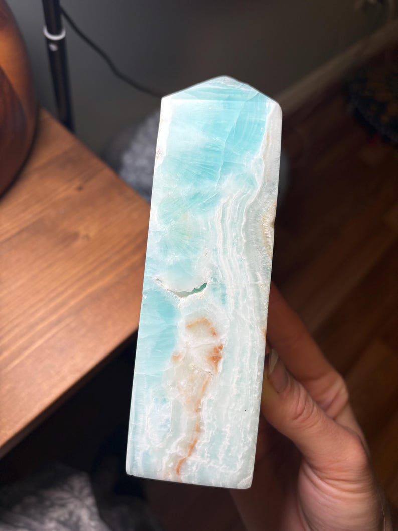May include: A polished tower of Caribbean calcite featuring a vibrant light blue color with white, wavy, translucent banding and small patches of orange-brown mineral inclusions throughout the stone's smooth, vertical, and rectangular structure.