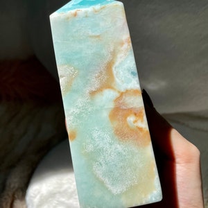 May include: A polished stone tower with a pointed top, featuring a smooth surface with a blend of light blue, teal, and warm tan or brown swirling patterns throughout the mineral structure, showcasing natural variations in color and texture.