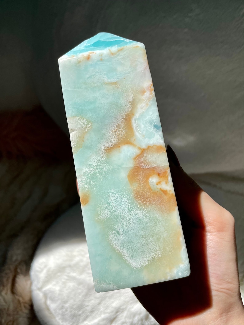 May include: A polished stone tower with a pointed top, featuring a smooth surface with a blend of light blue, teal, and warm tan or brown swirling patterns throughout the mineral structure, showcasing natural variations in color and texture.
