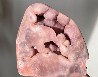 May include: A large, pink amethyst geode with a natural, rough exterior and a hollow interior filled with smaller, purple crystals. The geode is illuminated by sunlight, highlighting the intricate details of the crystals.