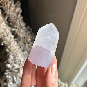 May include: A clear, faceted crystal tower with a soft lavender base. The crystal is held in a hand, with dried plants in the background. The crystal's shape is a rectangular prism with a pointed top, and it appears to be translucent.