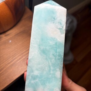 May include: A polished, tower-shaped crystal point featuring a smooth, multifaceted surface with a marbled appearance. The stone displays a vibrant blend of translucent light blue and opaque white colors, with a pointed top and a flat base.
