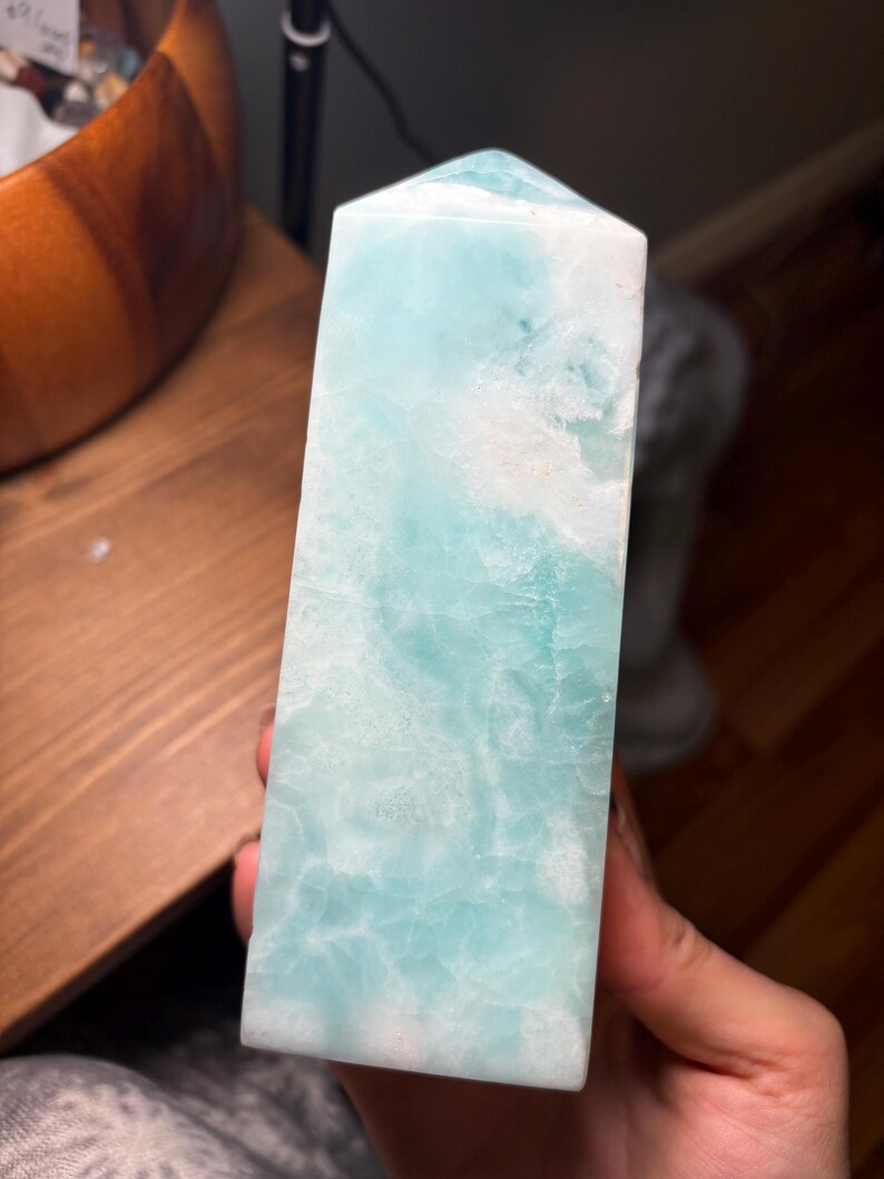 May include: A polished, tower-shaped crystal point featuring a smooth, multifaceted surface with a marbled appearance. The stone displays a vibrant blend of translucent light blue and opaque white colors, with a pointed top and a flat base.