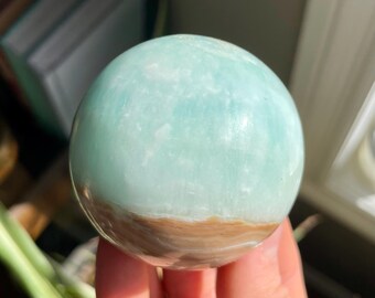 May include: A polished sphere with a light blue top and a brown bottom. The sphere is held in a hand, with a blurred background of plants and a window.