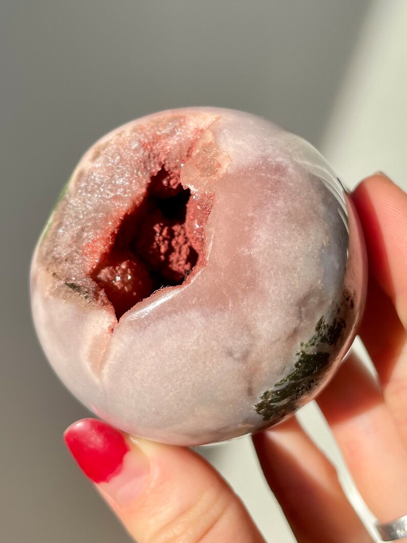 May include: A pink and green geode sphere with a large opening revealing a red and pink crystal inside.