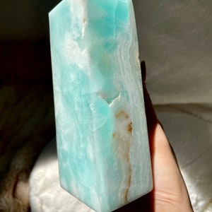 May include: A polished, tower-shaped crystal point in a vibrant shade of turquoise blue. The stone features a smooth, multifaceted surface with visible internal white veining and a distinct, narrow streak of earthy brown mineral inclusions.