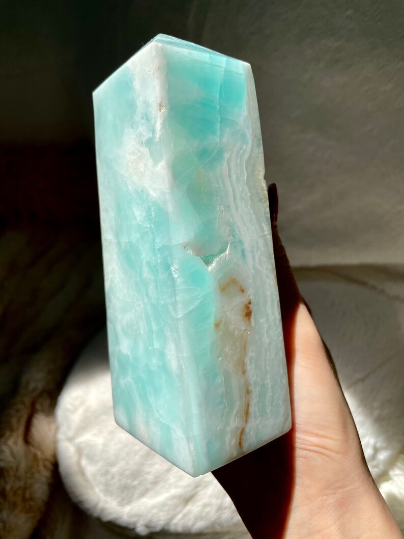 May include: A polished, tower-shaped crystal point in a vibrant shade of turquoise blue. The stone features a smooth, multifaceted surface with visible internal white veining and a distinct, narrow streak of earthy brown mineral inclusions.