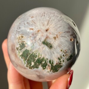 May include: A white and green agate sphere with a unique, swirling pattern. The sphere is polished and smooth, with a natural, organic look.