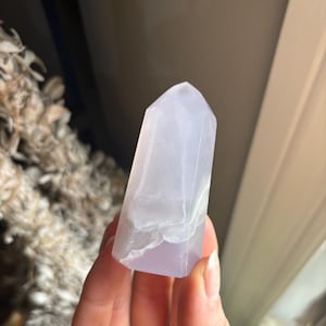 May include: A light purple crystal point with a faceted top. The crystal has a translucent appearance with white veining and is held in a hand. The crystal is a light purple colour.