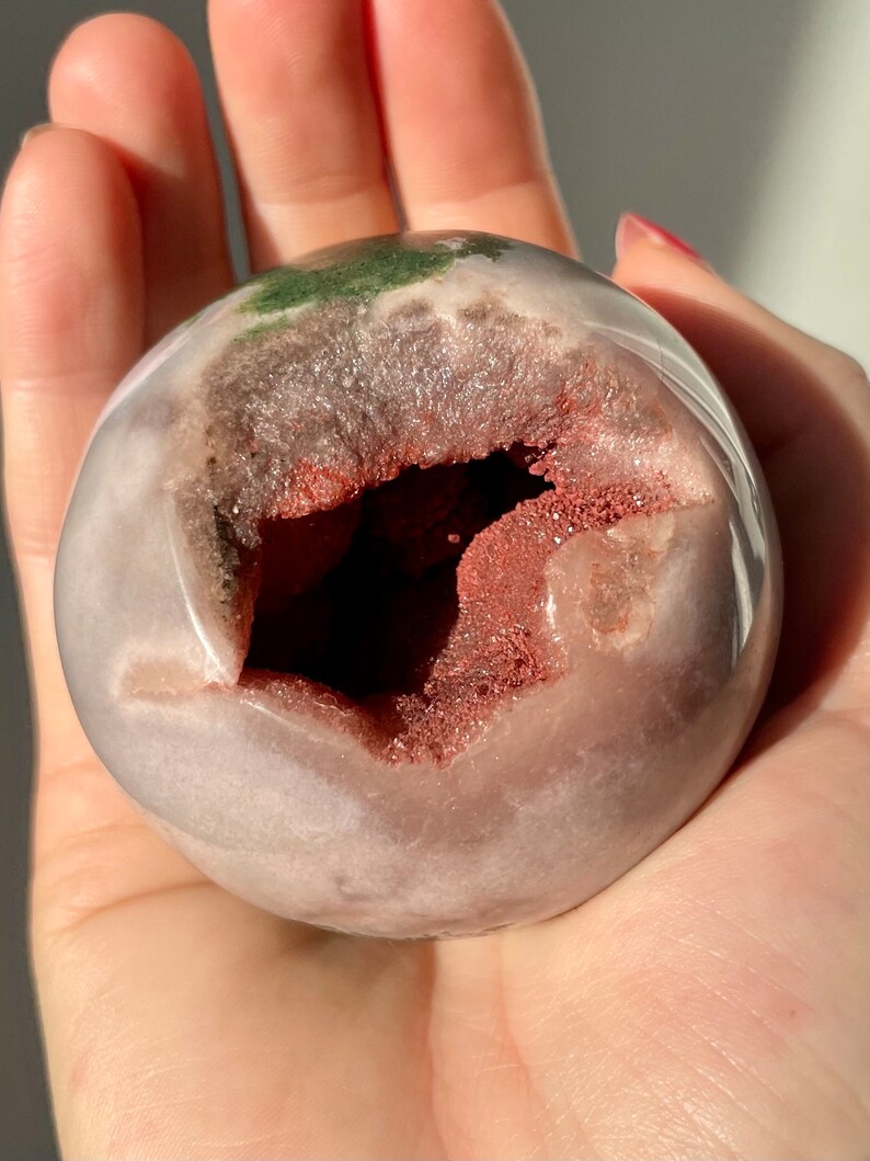 May include: A pink and white agate geode sphere with a large opening revealing a red and white crystal interior. The sphere is smooth and polished.