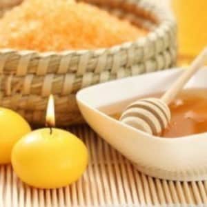 Puede incluir: Un bol blanco lleno de miel y un cucharón de miel de madera. Dos velas amarillas están encendidas en el fondo. También se puede ver una cesta de mimbre llena de sales de baño naranjas.