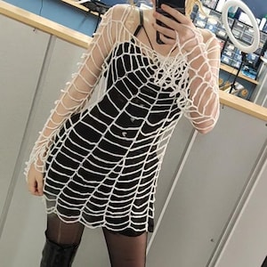 Może przedstawiać: Czarna sukienka pokryta jest białym, przypominającym pajęczynę wzorem z szydełka. Strój uzupełniają czarne buty na platformie. Ogólna estetyka sugeruje gotycki lub alternatywny styl.