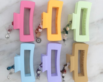 May include: Six colorful plastic hair clips with dangling charms. The charms are made of beads, stones, and metal. The clips are in the colors pink, yellow, light green, light blue, purple, and brown.