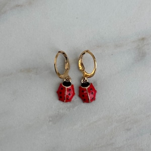 Puede incluir: Un par de pendientes de aro dorados con colgantes de mariquita rojos. Cada colgante de mariquita tiene lunares negros y una cabeza negra. Los pendientes se exhiben sobre una superficie de mármol blanco y gris.