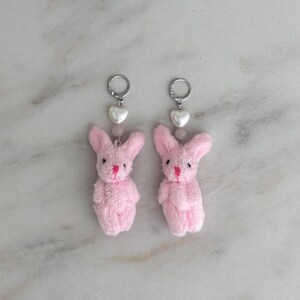 May include: A pair of pink bunny rabbit charm earrings with a white heart charm and silver hoop.