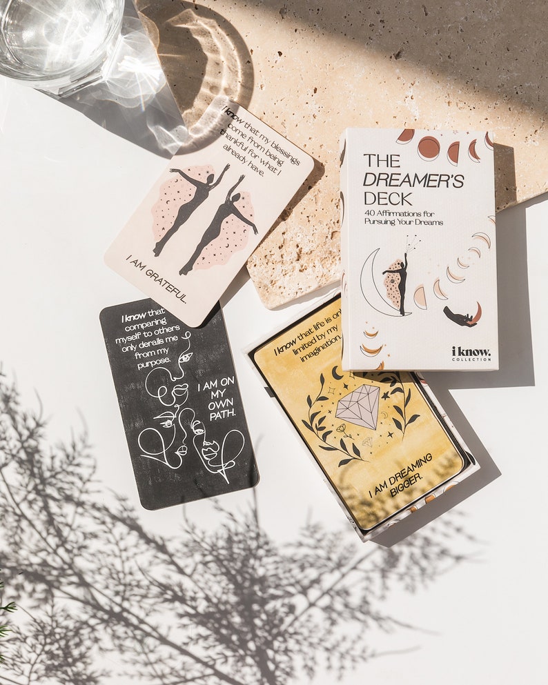 Puede incluir: Un juego de cartas de afirmaci&oacute;n con ilustraciones y texto. Las cartas son beige, negras y amarillas con fondo blanco. El texto en las cartas incluye "Estoy agradecida", "Estoy en mi propio camino", "S&eacute; que la vida solo est&aacute; limitada por mi imaginaci&oacute;n", "Estoy so&ntilde;ando m&aacute;s grande", y "La baraja del so&ntilde;ador 40 afirmaciones para perseguir tus sue&ntilde;os".