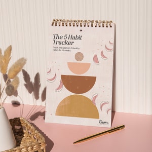 May include: A spiral bound journal with a gold wire binding and a beige cover. The cover features a geometric design with pink, brown, and gold shapes. The text "The 5 Habit Tracker" is printed in black at the top of the cover. The text "Track and Maintain 5 Healthy Habits for 16-weeks" is printed in black below the title. The text "the know." is printed in black at the bottom of the cover.