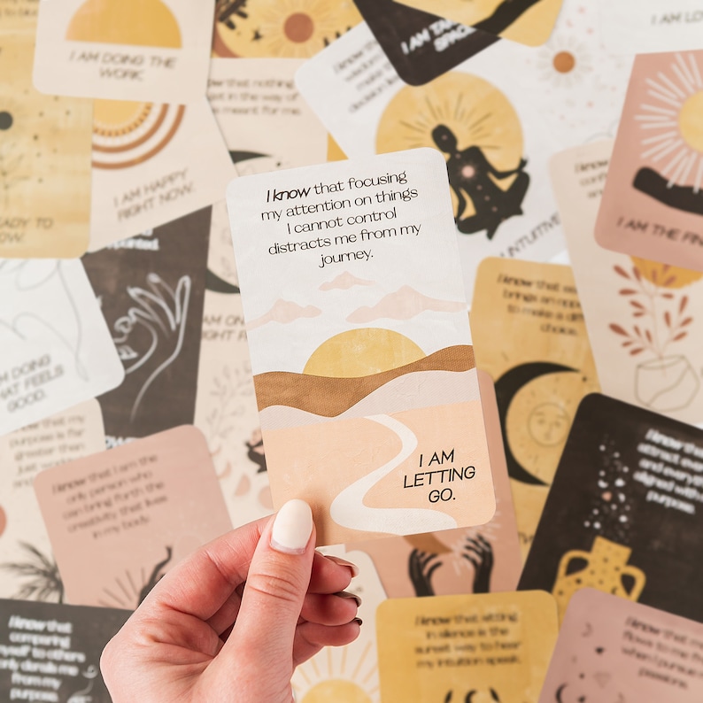 Puede incluir: Un juego de cartas de afirmaci&oacute;n con una mano sosteniendo una carta que dice "Estoy dejando ir". La carta tiene un dise&ntilde;o de puesta de sol y un camino sinuoso. Las otras cartas tienen varios dise&ntilde;os y afirmaciones.