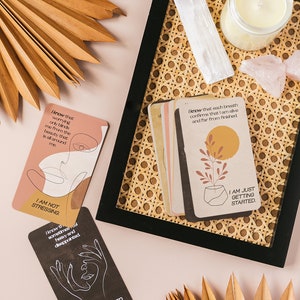 Puede incluir: Un juego de cartas de afirmaci&oacute;n con mensajes positivos e ilustraciones. Las cartas est&aacute;n en una bandeja tejida con una vela, cristales y hojas de palma secas.