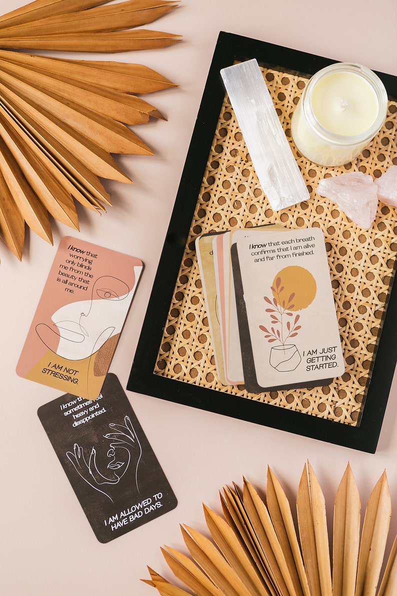 Puede incluir: Un juego de cartas de afirmaci&oacute;n con mensajes positivos e ilustraciones. Las cartas est&aacute;n en una bandeja tejida con una vela, cristales y hojas de palma secas.