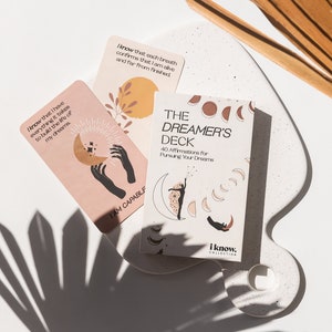 Puede incluir: Una baraja de cartas de afirmaci&oacute;n llamada "The Dreamer's Deck" con 40 afirmaciones para perseguir tus sue&ntilde;os. Las cartas presentan un dise&ntilde;o de luna y estrellas con una mujer con un vestido fluido de pie sobre una luna creciente. El texto en la parte delantera de la baraja dice "The Dreamer's Deck 40 Affirmations for Pursuing Your Dreams".