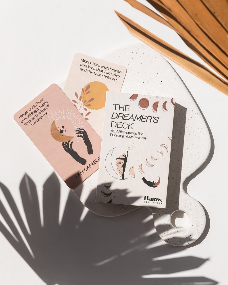 Puede incluir: Una baraja de cartas de afirmaci&oacute;n llamada "The Dreamer's Deck" con 40 afirmaciones para perseguir tus sue&ntilde;os. Las cartas presentan un dise&ntilde;o de luna y estrellas con una mujer con un vestido fluido de pie sobre una luna creciente. El texto en la parte delantera de la baraja dice "The Dreamer's Deck 40 Affirmations for Pursuing Your Dreams".