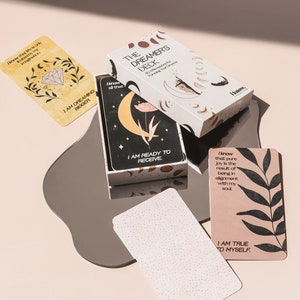 Puede incluir: Una caja blanca con el texto "The Dreamer's Deck 40 Affirmations for Nurturing Your Dreams" en la parte delantera. La caja est&aacute; abierta y muestra una tarjeta con el texto "I am ready to receive" y una luna creciente con una flor. La tarjeta est&aacute; sobre una superficie plateada con una tarjeta blanca con el texto "I am true to myself" sobre una superficie rosa.