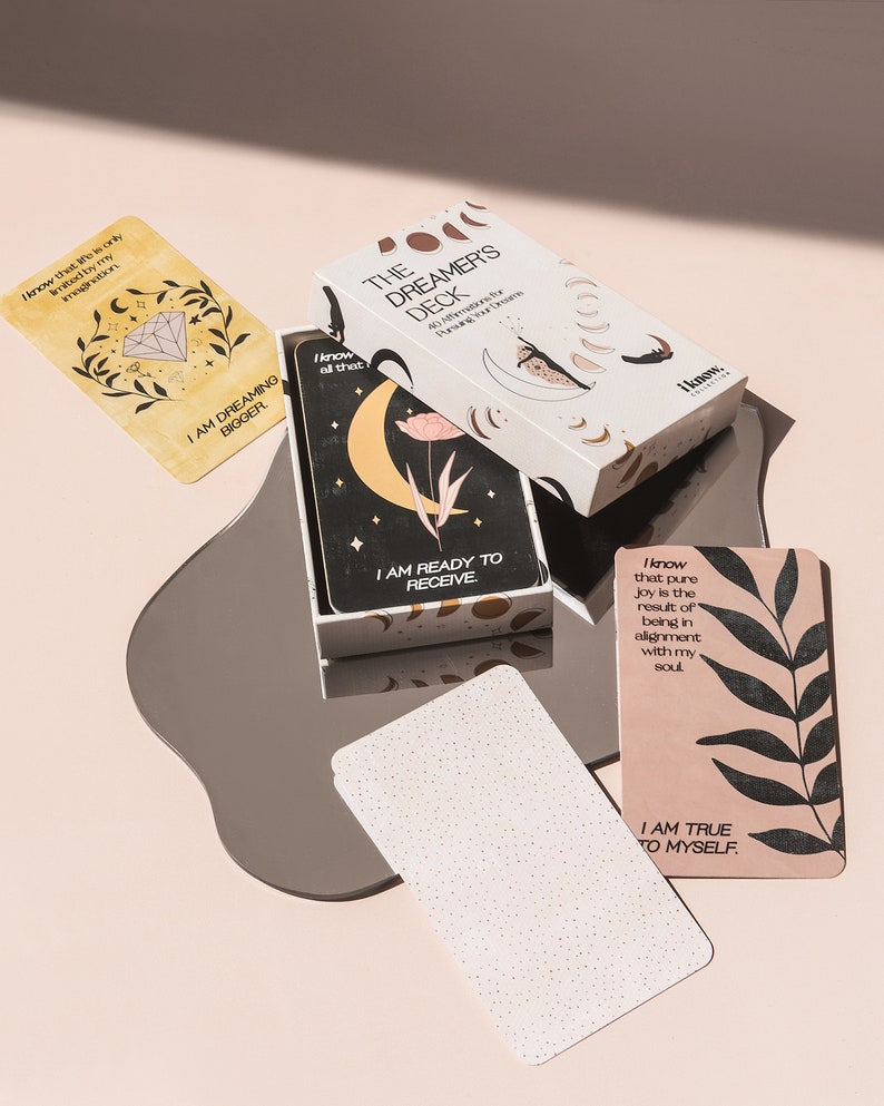 Puede incluir: Una caja blanca con el texto "The Dreamer's Deck 40 Affirmations for Nurturing Your Dreams" en la parte delantera. La caja est&aacute; abierta y muestra una tarjeta con el texto "I am ready to receive" y una luna creciente con una flor. La tarjeta est&aacute; sobre una superficie plateada con una tarjeta blanca con el texto "I am true to myself" sobre una superficie rosa.