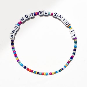 以下が含まれることがあります： 白い文字ビーズで「AND NOW WE SAID IT」と綴られた、カラフルなビーズブレスレットです。