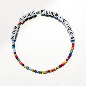 以下が含まれることがあります： 白い文字ビーズで「She's Pernicious」と「Wow」と綴られた、カラフルなビーズブレスレットです。