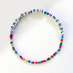 Puede incluir: Un brazalete de cuentas de colores con cuentas de letras blancas que deletrean "Don't Be All Uncool".