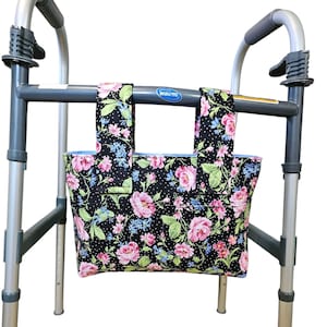 May include: A black walker bag with a floral pattern of pink roses, blue flowers, and green leaves. The bag is attached to a gray walker frame. The bag has a light blue interior and is designed to hold personal items.