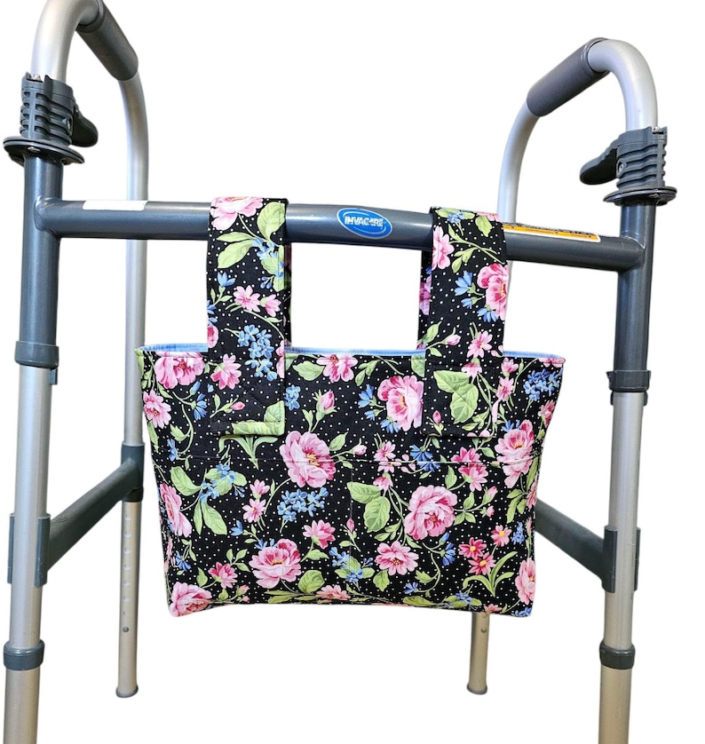 Puede incluir: Una bolsa negra para andador con un estampado floral de rosas rosas, flores azules y hojas verdes. La bolsa est&aacute; sujeta a un marco de andador gris. La bolsa tiene un interior azul claro y est&aacute; dise&ntilde;ada para guardar objetos personales.