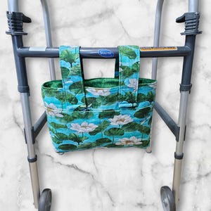 May include: A fabric walker bag with a floral pattern of white lotus flowers and green leaves on a turquoise background. The bag is attached to a gray walker frame, providing a convenient storage solution for mobility aids.