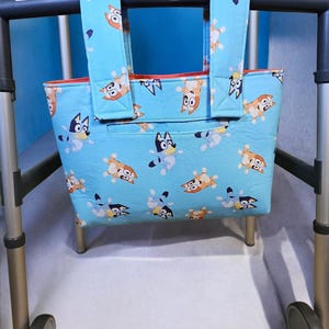 May include: A light blue tote bag with two matching fabric handles, featuring a repeating pattern of cartoon dogs in various poses. The bag is attached to the horizontal frame of a metal walker, which has a blue Invacare label on the top bar.