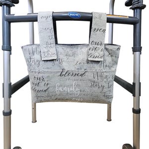 May include: A grey walker with a fabric bag attached. The bag is light grey with white script that reads "blessed", "family", and other words. The walker has black handles and silver legs and wheels.